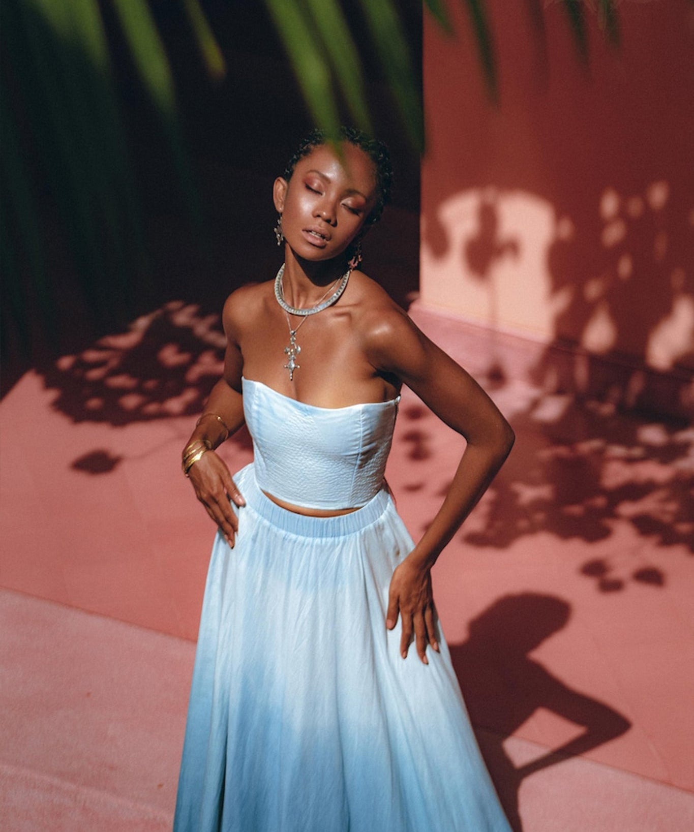A woman stands gracefully in the Boho Top Corset Skirt Set by AYA Sacred Wear, eyes closed, as sunlight casts leaf shadows around her. The backdrop of a pinkish wall and green foliage above enhances the serene, dreamy atmosphere.