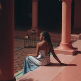 A woman with long braided hair sits on a pink ledge in front of a tiled floor. She wears the Boho Top Corset Skirt Set from AYA Sacred Wear and faces away from the camera. Lanterns and columns surround her, while sunlight and shadows create patterns on the ground.