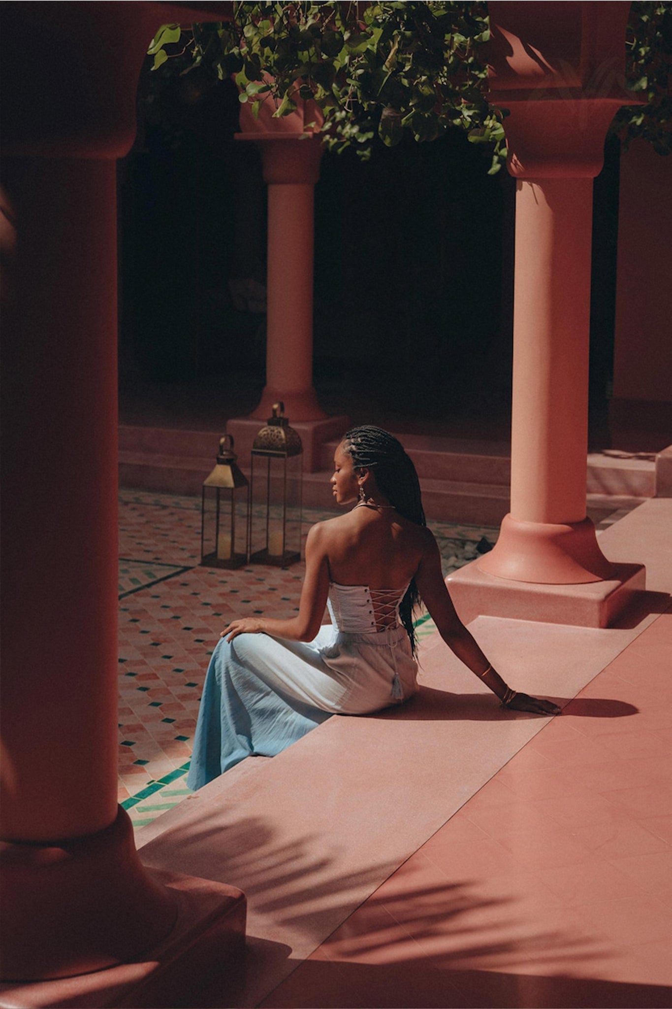 A woman with long braided hair sits on a pink ledge in front of a tiled floor. She wears the Boho Top Corset Skirt Set from AYA Sacred Wear and faces away from the camera. Lanterns and columns surround her, while sunlight and shadows create patterns on the ground.