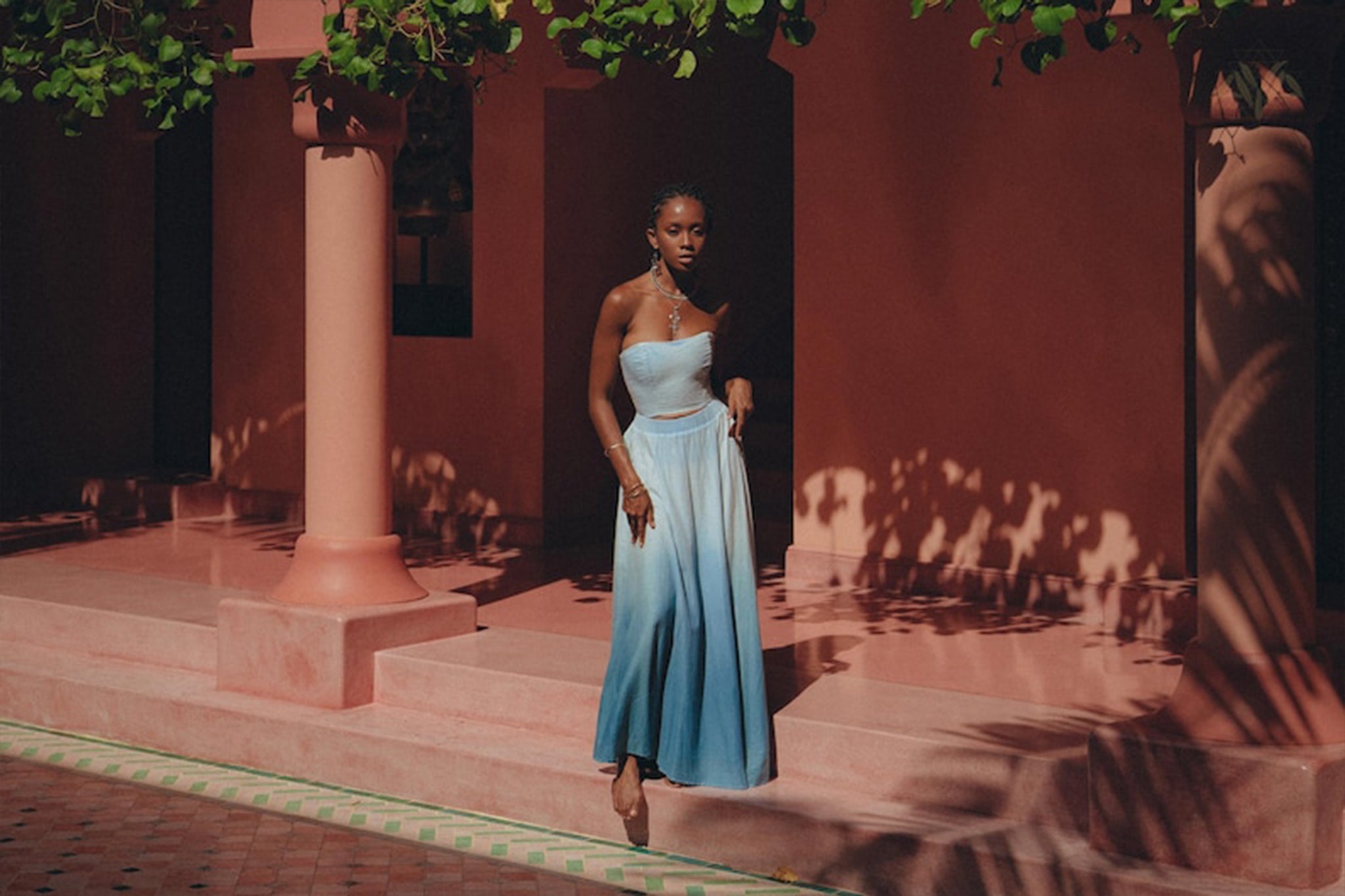 A woman wearing the AYA Sacred Wear Boho Top Corset Skirt Set, a botanically dyed set featuring a strapless blue ombre design with a flowing skirt, stands elegantly on pink steps before a building with coral pillars. Sunlight filters through nearby foliage, casting intricate shadows on the walls and ground.