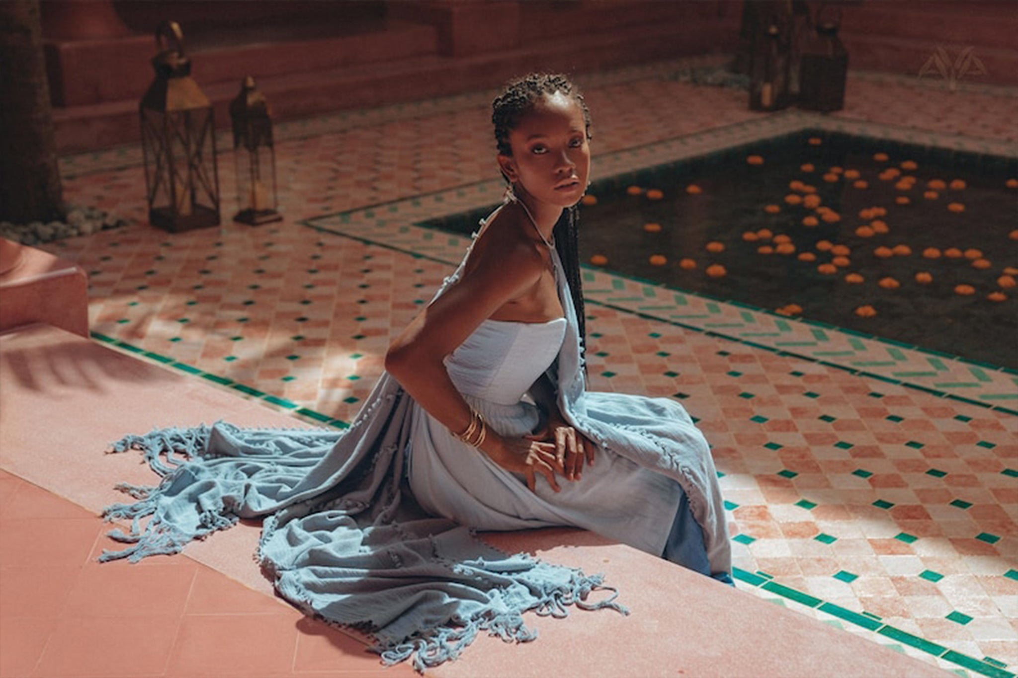 A person dressed in an AYA Sacred Wear Boho Top Corset Skirt Set sits gracefully on the edge of a decorative tiled pool filled with floating oranges. The scene is warmly lit, creating a serene and elegant atmosphere.