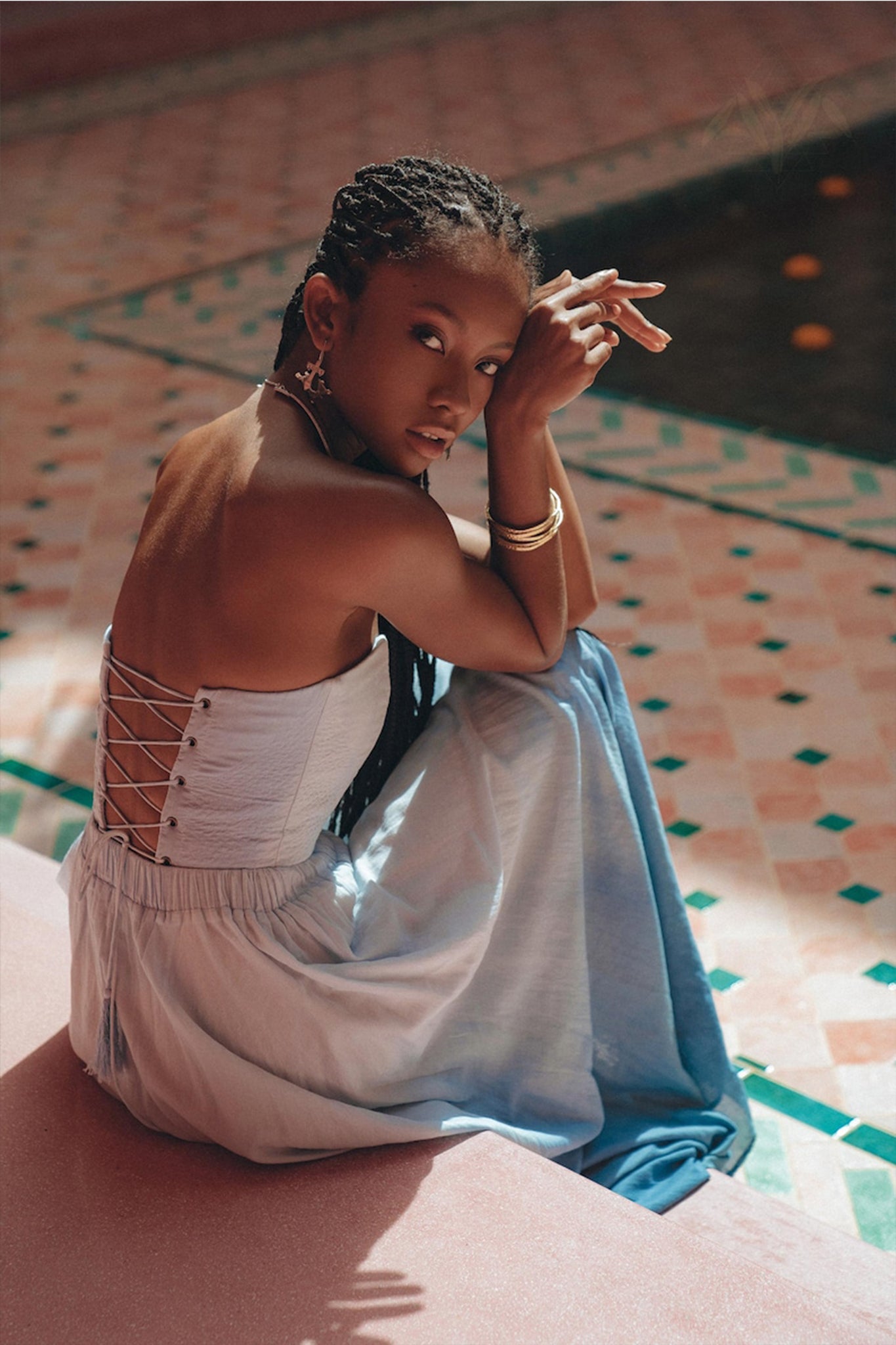 A person with long braided hair sits beside a tiled pool, wearing the Boho Top Corset Skirt Set from AYA Sacred Wear, which features a strapless design with a corset-style lace-up back. They accessorize with hoop earrings and rest an elbow on their knee, gazing into the distance while surrounded by a patterned tile floor.