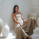 A woman sits gracefully against a white wall, surrounded by substantial ceramic vases. She is adorned in the Sage Green Summer Crop Top Skirt set from AYA Sacred Wear, featuring a flowing design that exudes elegance. Her long hair cascades loosely around her shoulders. The minimalist and tranquil setting accentuates neutral tones and textures, enhanced by hand-painted details that impart an artistic flair.