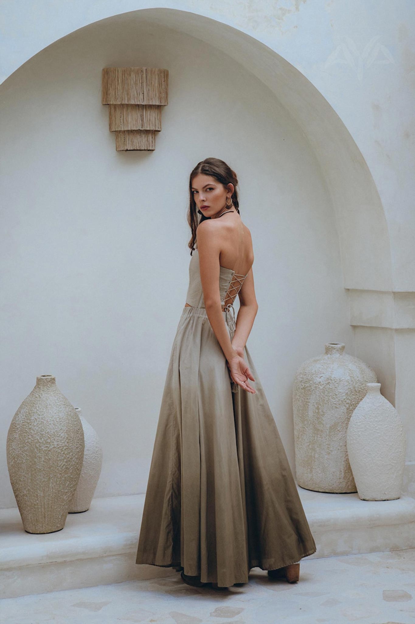 A woman wearing the Boho Top Corset and Skirt Women set by AYA Sacred Wear stands in a minimalist, arched alcove. She is surrounded by textured, neutral-toned pottery, creating an elegant and serene atmosphere.