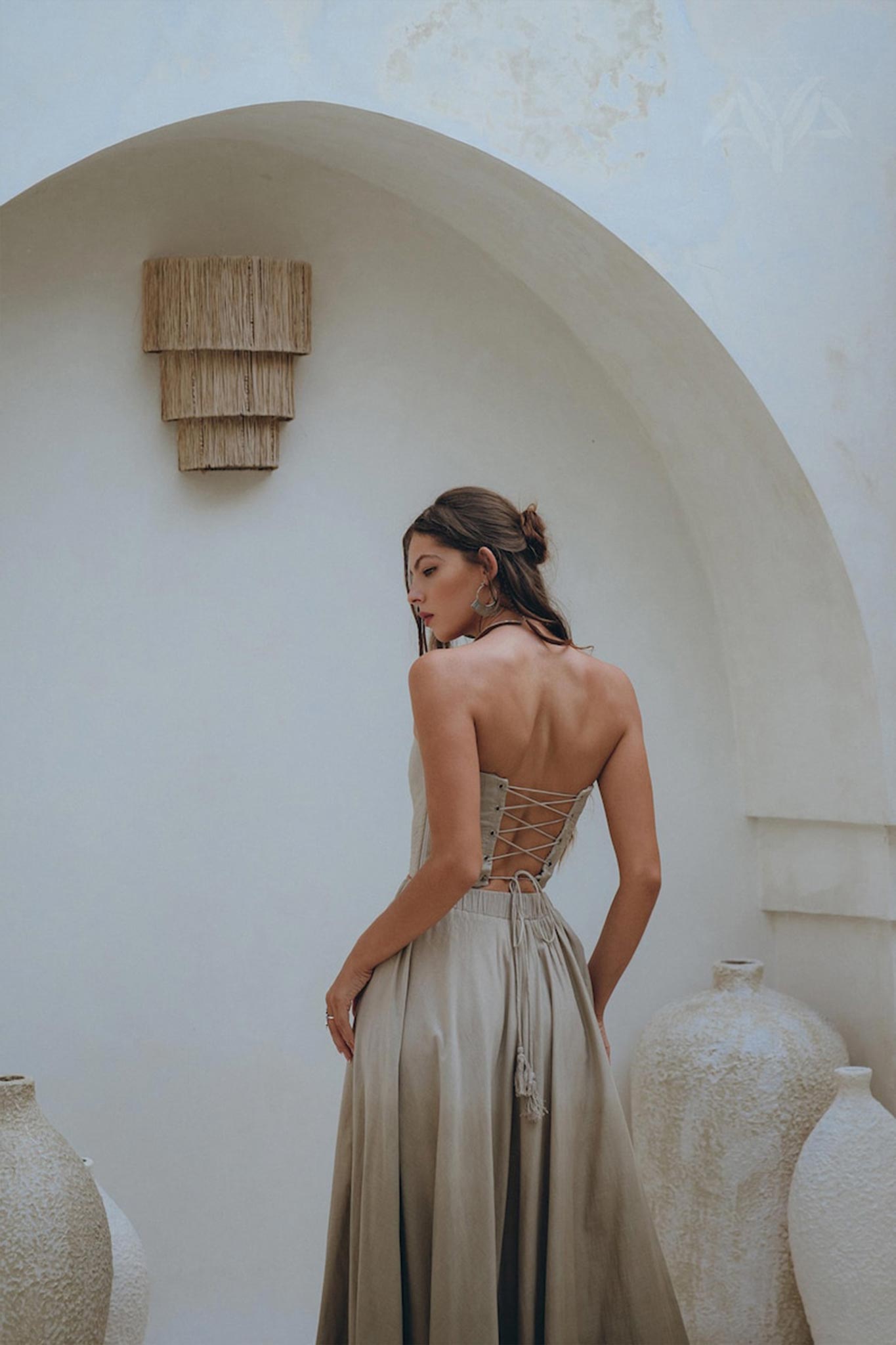 A woman in a sage green summer crop top and skirt from AYA Sacred Wear, featuring a boho-style corset design, stands near large ceramic vases against a textured white archway. Her hair is styled in a loose bun, and she gazes to the side, creating a serene and elegant atmosphere.