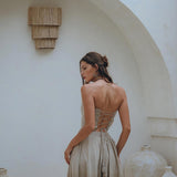 A woman in a sage green summer crop top and skirt from AYA Sacred Wear, featuring a boho-style corset design, stands near large ceramic vases against a textured white archway. Her hair is styled in a loose bun, and she gazes to the side, creating a serene and elegant atmosphere.