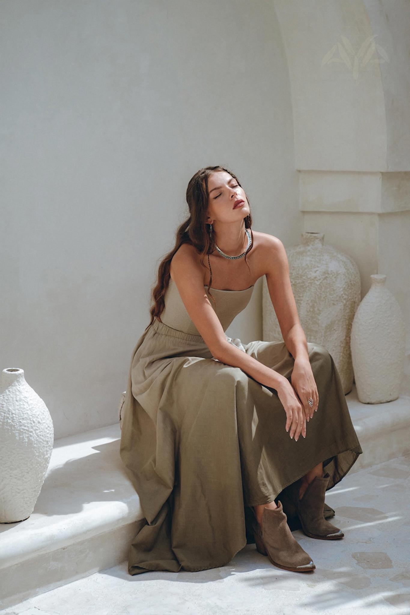 A woman with long hair sits on a white ledge, dressed in the Boho Top Corset and Skirt Women by AYA Sacred Wear. She wears the Sage Green Summer Crop Top Skirt amidst hand-painted white ceramic vases, with natural sunlight creating a calm and relaxed atmosphere around her.