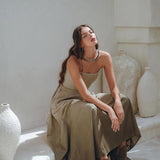 A woman with long hair sits on a white ledge, dressed in the Boho Top Corset and Skirt Women by AYA Sacred Wear. She wears the Sage Green Summer Crop Top Skirt amidst hand-painted white ceramic vases, with natural sunlight creating a calm and relaxed atmosphere around her.