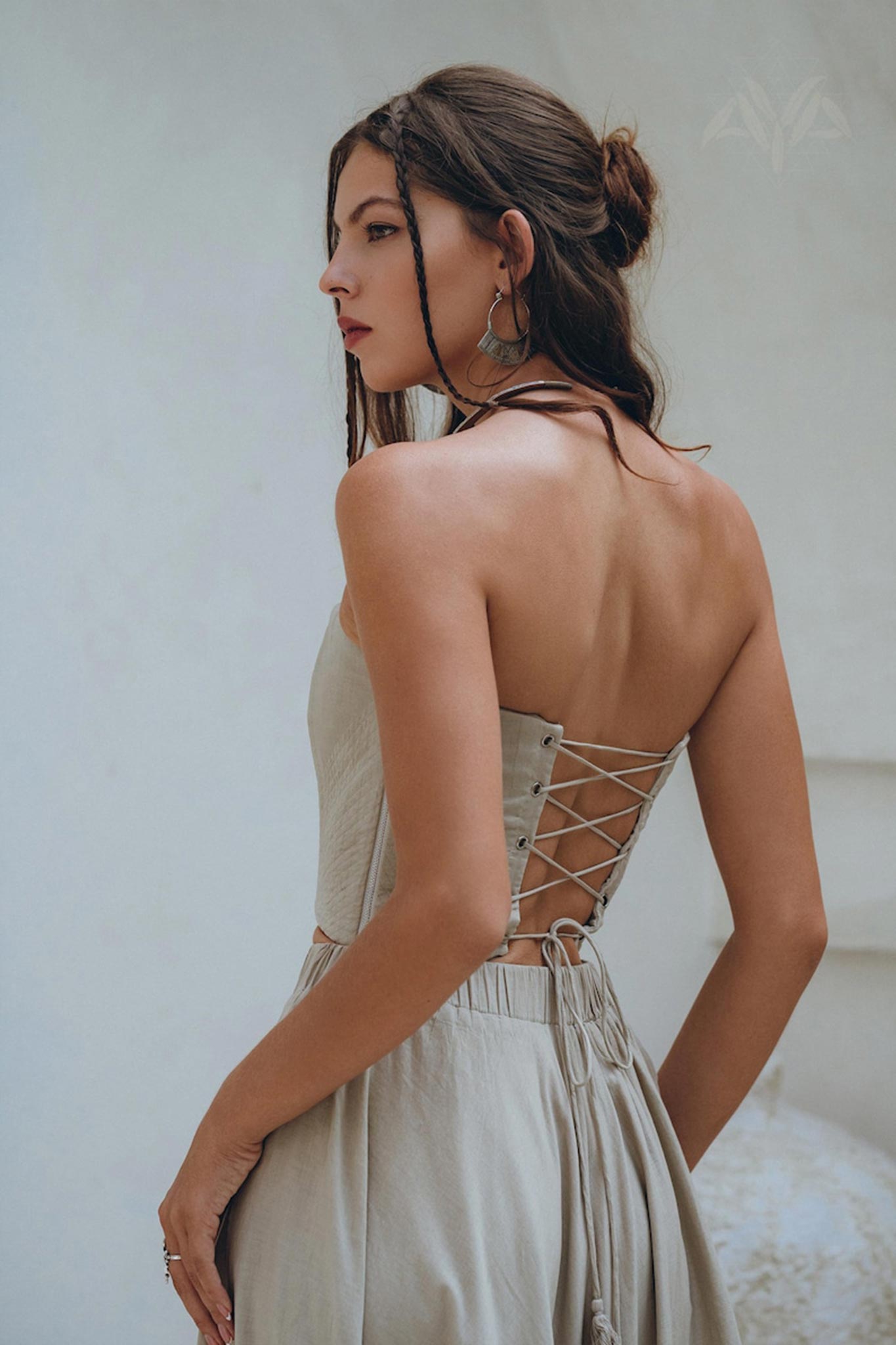 A woman with long braided hair elegantly wears the Boho Top Corset and Skirt Women in a sage green shade, a summer crop top and skirt set from AYA Sacred Wear. The lace-up back corset design of the dress offers a unique blend of style and sustainability, creating an eye-catching contrast against the plain background.