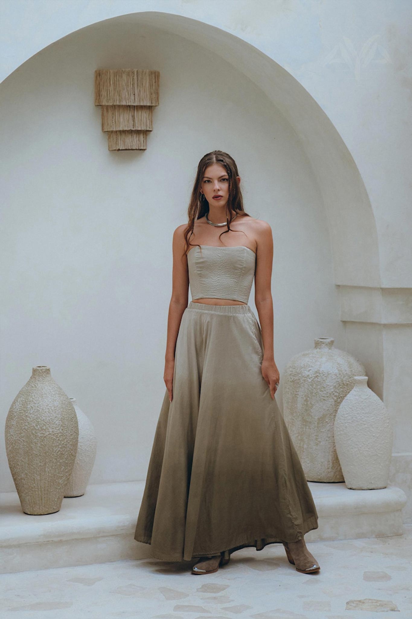 A woman poses gracefully in the Boho Top Corset and Skirt by AYA Sacred Wear, showcasing the sage green summer crop top and skirt set against a minimalist backdrop featuring white textured vases and a curved arch, creating a soft, elegant atmosphere.