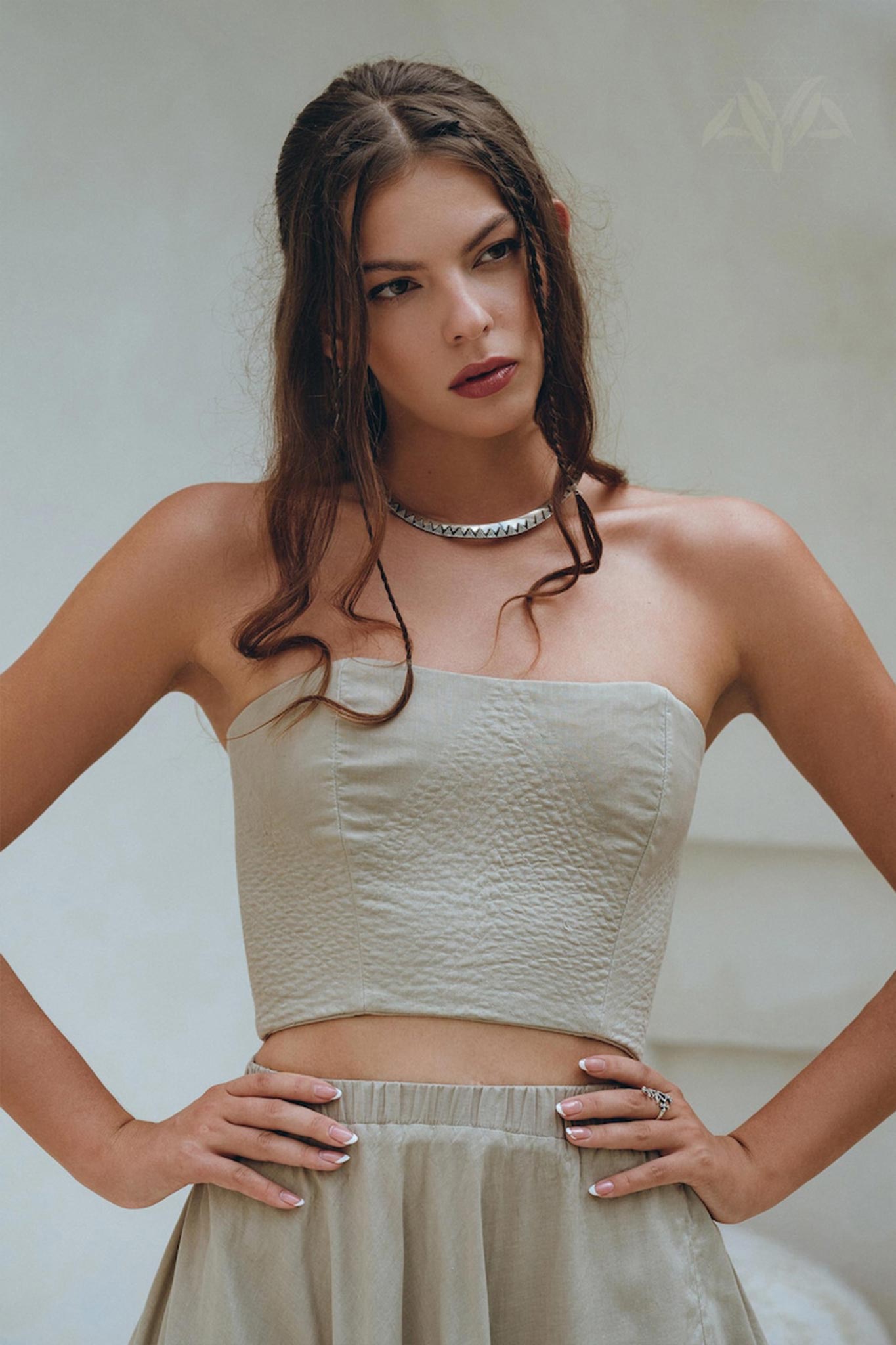 A woman with long brown hair stands looking to the side, wearing AYA Sacred Wear's Boho Top Corset and Skirt in a sage green hue. She accessorizes with a silver necklace and ring, her hands confidently on her hips against a neutral background.