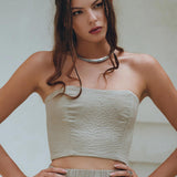 A woman with long brown hair stands looking to the side, wearing AYA Sacred Wear's Boho Top Corset and Skirt in a sage green hue. She accessorizes with a silver necklace and ring, her hands confidently on her hips against a neutral background.