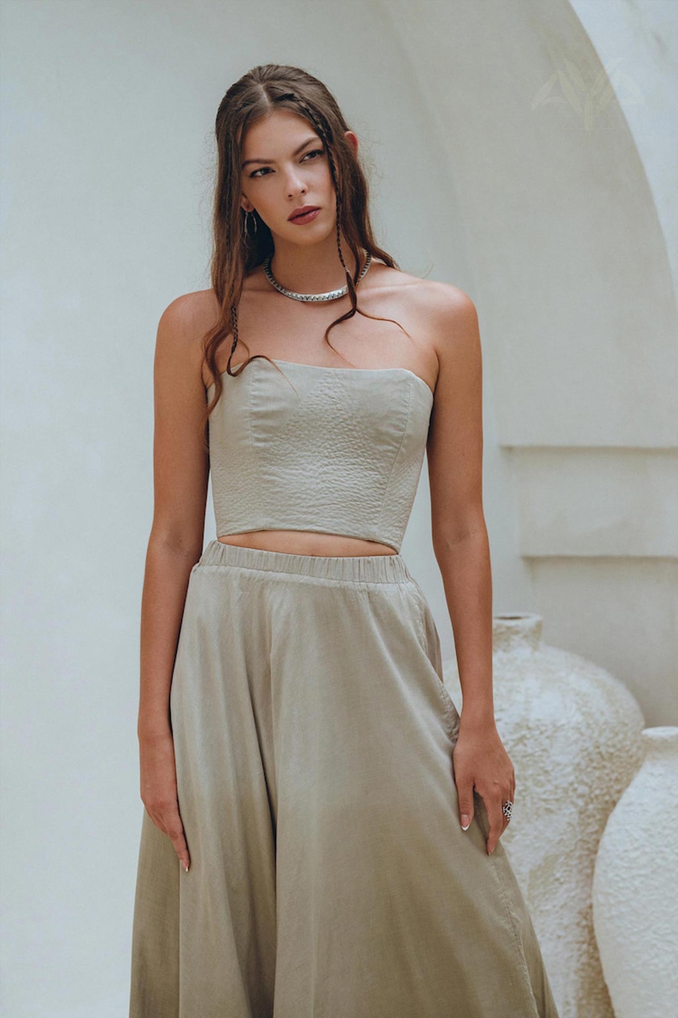 In a serene pose, a woman is against a white backdrop, dressed in an AYA Sacred Wear sage green summer crop top and skirt. Her wavy hair cascades gracefully over her shoulders, and she wears a calm expression accented by a necklace. Large vases are part of the scene behind her.