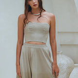 In a serene pose, a woman is against a white backdrop, dressed in an AYA Sacred Wear sage green summer crop top and skirt. Her wavy hair cascades gracefully over her shoulders, and she wears a calm expression accented by a necklace. Large vases are part of the scene behind her.