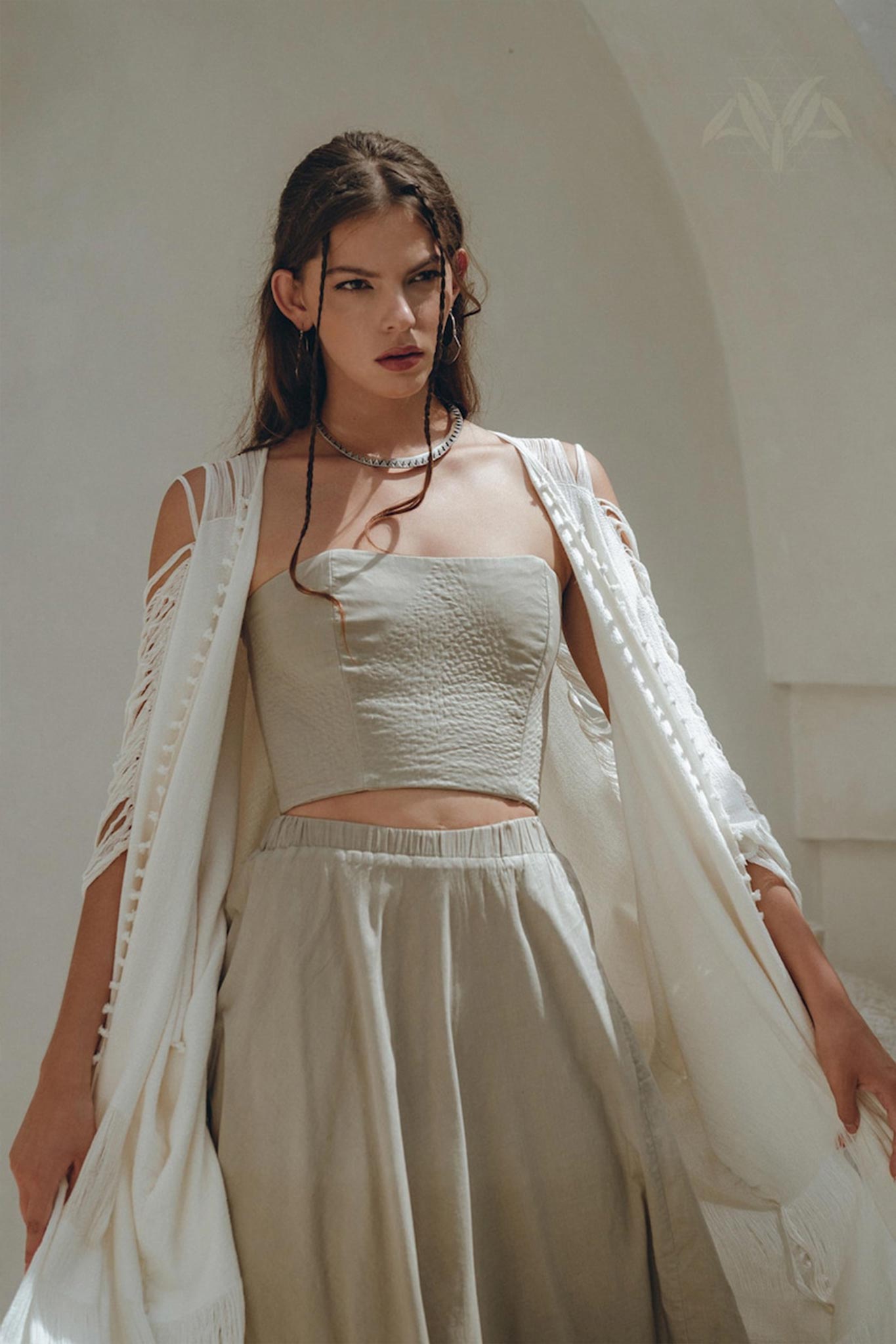 A woman in a sage green, sleeveless corset top and matching boho skirt from AYA Sacred Wear poses outdoors. Her light, loose white shawl drapes over her shoulders, complementing her slightly wavy hair. The backdrop of a soft, neutral-toned wall adds to the serene setting.