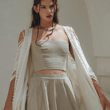 A woman in a sage green, sleeveless corset top and matching boho skirt from AYA Sacred Wear poses outdoors. Her light, loose white shawl drapes over her shoulders, complementing her slightly wavy hair. The backdrop of a soft, neutral-toned wall adds to the serene setting.