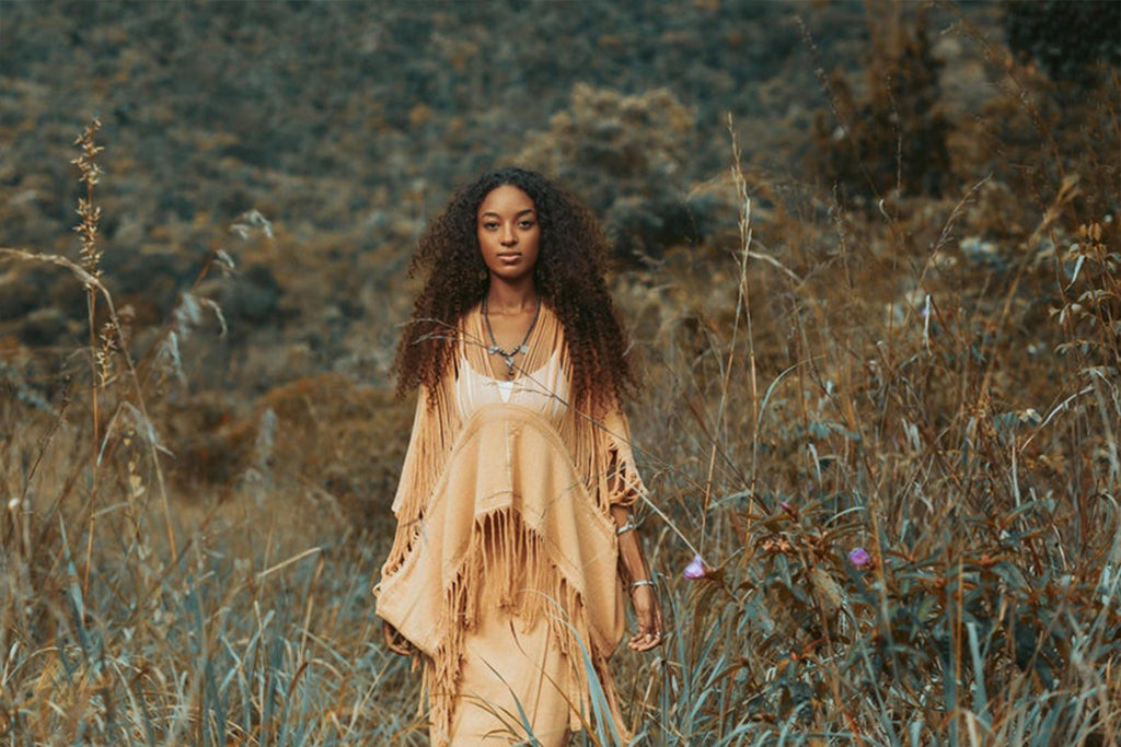 A woman with long curly hair, dressed in a tan shawl and dress complemented by AYA Sacred Wear's Boho Top, elegantly crafted from hand-loomed cotton, strolls through a field of tall grasses. The backdrop reveals a hillside adorned with muted green and brown vegetation, crafting a serene and natural ambiance.