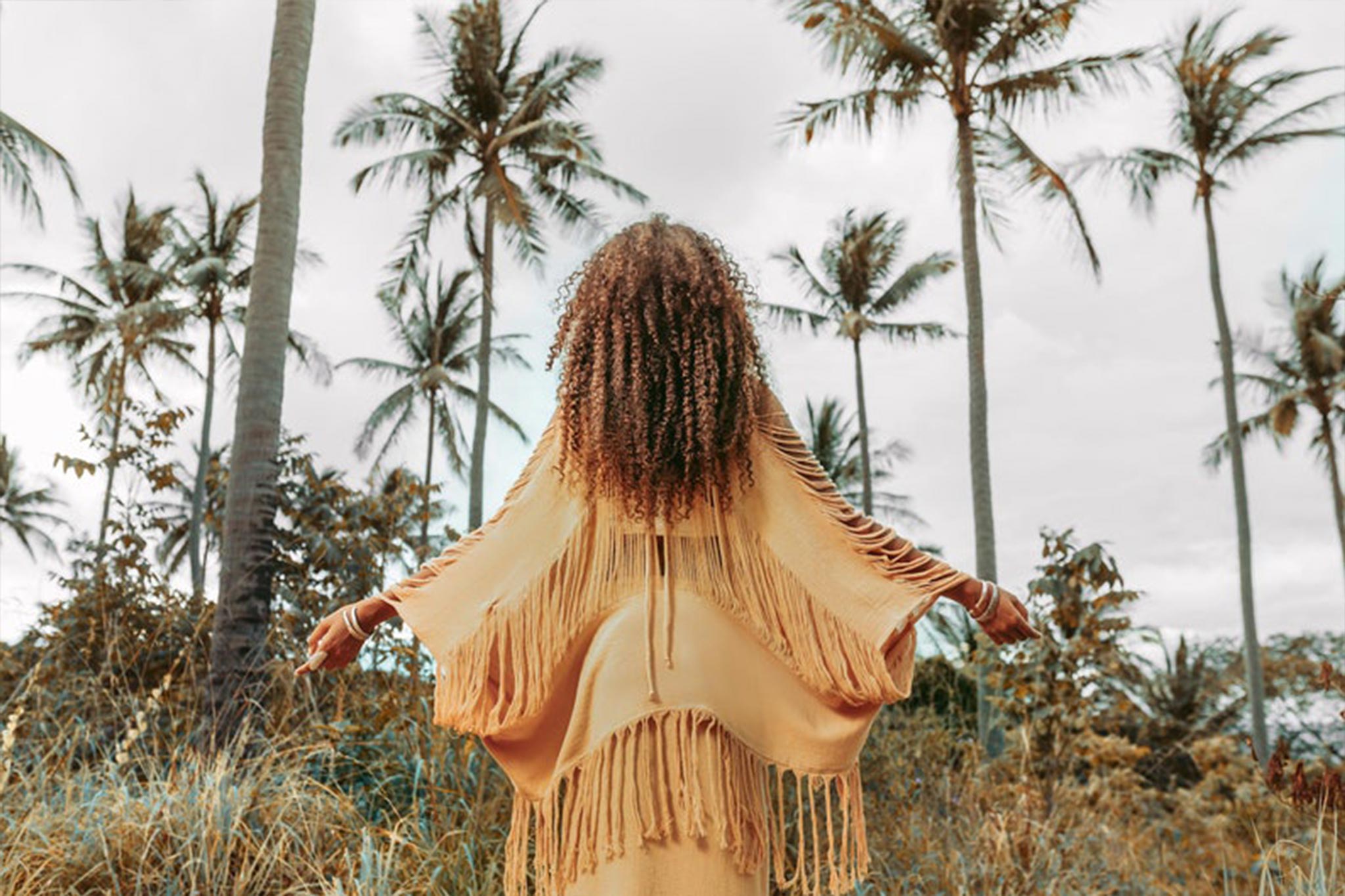 A person with curly hair, adorned in an AYA Sacred Wear Boho Top—a hand loomed cotton blouse—stands amidst tall palm trees, their fringed shawl flowing gently. The overcast sky embraces the tranquil essence of nature.