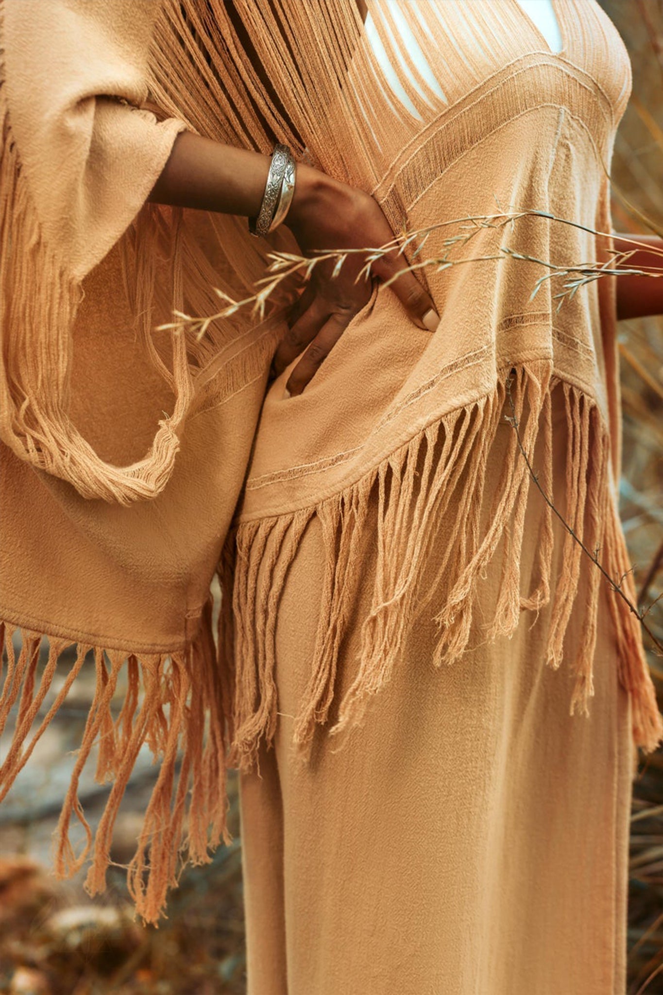 A person wearing the AYA Sacred Wear Boho Top, a beige hand-loomed cotton blouse with bohemian fringe details, stands with hands on hips. A silver bracelet adorns their wrist. Dry grass in the background enhances the warm, earthy ambiance that evokes sustainable elegance.