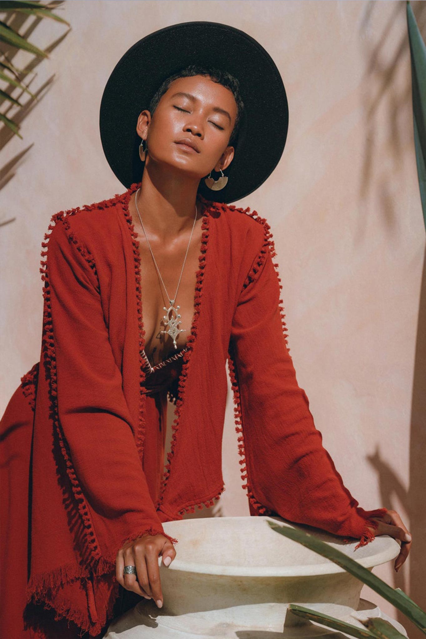 A person leans against a white stone planter, eyes closed. They are adorned in a red textured shawl draped over an AYA Sacred Wear's Boho Top—an Organic Wide Sleeve Wrap Top for women. Topping their look is a black wide-brimmed hat and large silver hoop earrings, with the sunlit wall behind casting partial shadows of foliage.