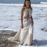 A woman stands gracefully in shallow ocean water, adorned in a Boho Wedding Dress from AYA Sacred Wear, featuring an open back and sides belly design with handwoven fabric. The sky is partly cloudy, and waves gently crash around her feet in a serene coastal setting reminiscent of Ayurvedic harmony with nature.