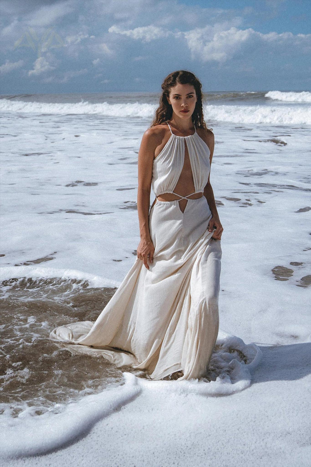 A woman stands gracefully in shallow ocean water, adorned in a Boho Wedding Dress from AYA Sacred Wear, featuring an open back and sides belly design with handwoven fabric. The sky is partly cloudy, and waves gently crash around her feet in a serene coastal setting reminiscent of Ayurvedic harmony with nature.