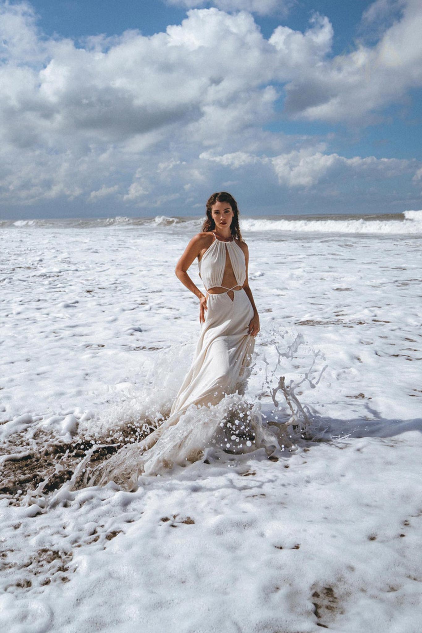 A woman in a Boho Wedding Dress for Women, an open-back sides belly dress designed by AYA Sacred Wear, stands on a beach, surrounded by foamy waves. The sky above is cloudy, and the ocean creates a dramatic backdrop.