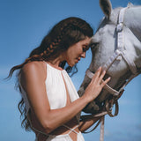 A woman wearing a Boho Wedding Dress for Women by AYA Sacred Wear, featuring an open back and sides, gently touches foreheads with a white horse on a sunny beach. The sky is clear and blue, creating a serene and intimate moment.