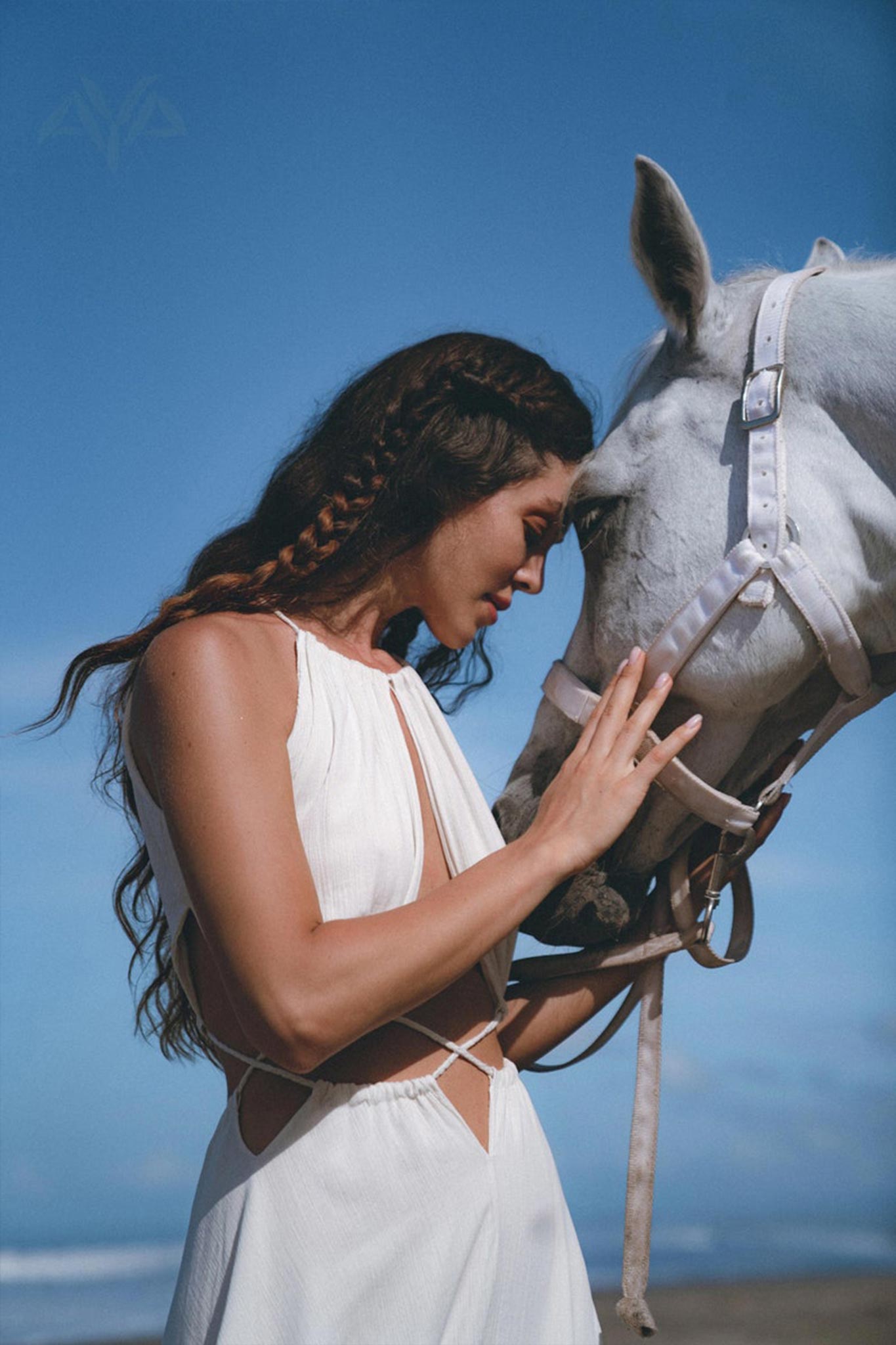 A woman wearing a Boho Wedding Dress for Women by AYA Sacred Wear, featuring an open back and sides, gently touches foreheads with a white horse on a sunny beach. The sky is clear and blue, creating a serene and intimate moment.