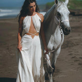 A woman wearing the Boho Wedding Dress for Women by AYA Sacred Wear, a stunning open-back design in flowing white, strolls barefoot along a beach, holding the reins of a white horse. Ocean waves crash majestically while lush greenery can be seen under the clear blue sky in the background.