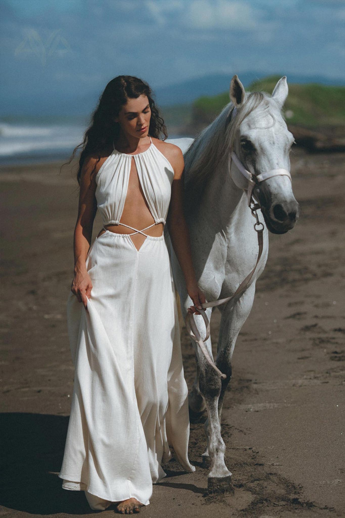 A woman wearing the Boho Wedding Dress for Women by AYA Sacred Wear, a stunning open-back design in flowing white, strolls barefoot along a beach, holding the reins of a white horse. Ocean waves crash majestically while lush greenery can be seen under the clear blue sky in the background.