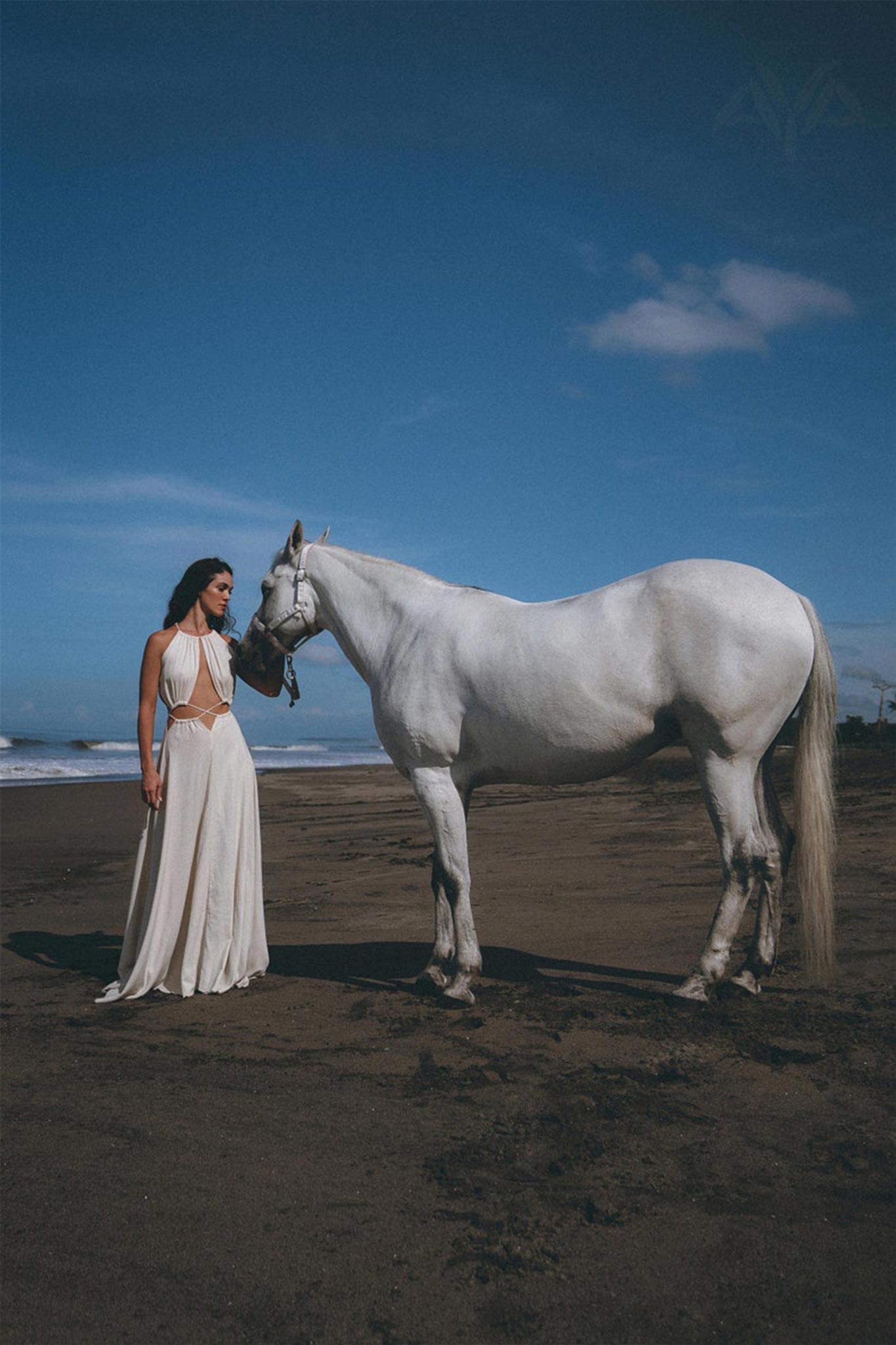 A woman in an AYA Sacred Wear Boho Wedding Dress for Women, featuring an open back and sides belly design, stands beside a white horse on a sandy beach beneath a clear blue sky.