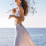 A woman stands by the sea wearing the Boho Wedding Dress for Women from AYA Sacred Wear, featuring an open back and sides-belly design. Made of organic botanically dyed fabric, the dress flows beautifully around her as she holds a delicate branch. The sun casts a warm glow on her as the breeze gently moves her hair, creating a serene and dreamy atmosphere.