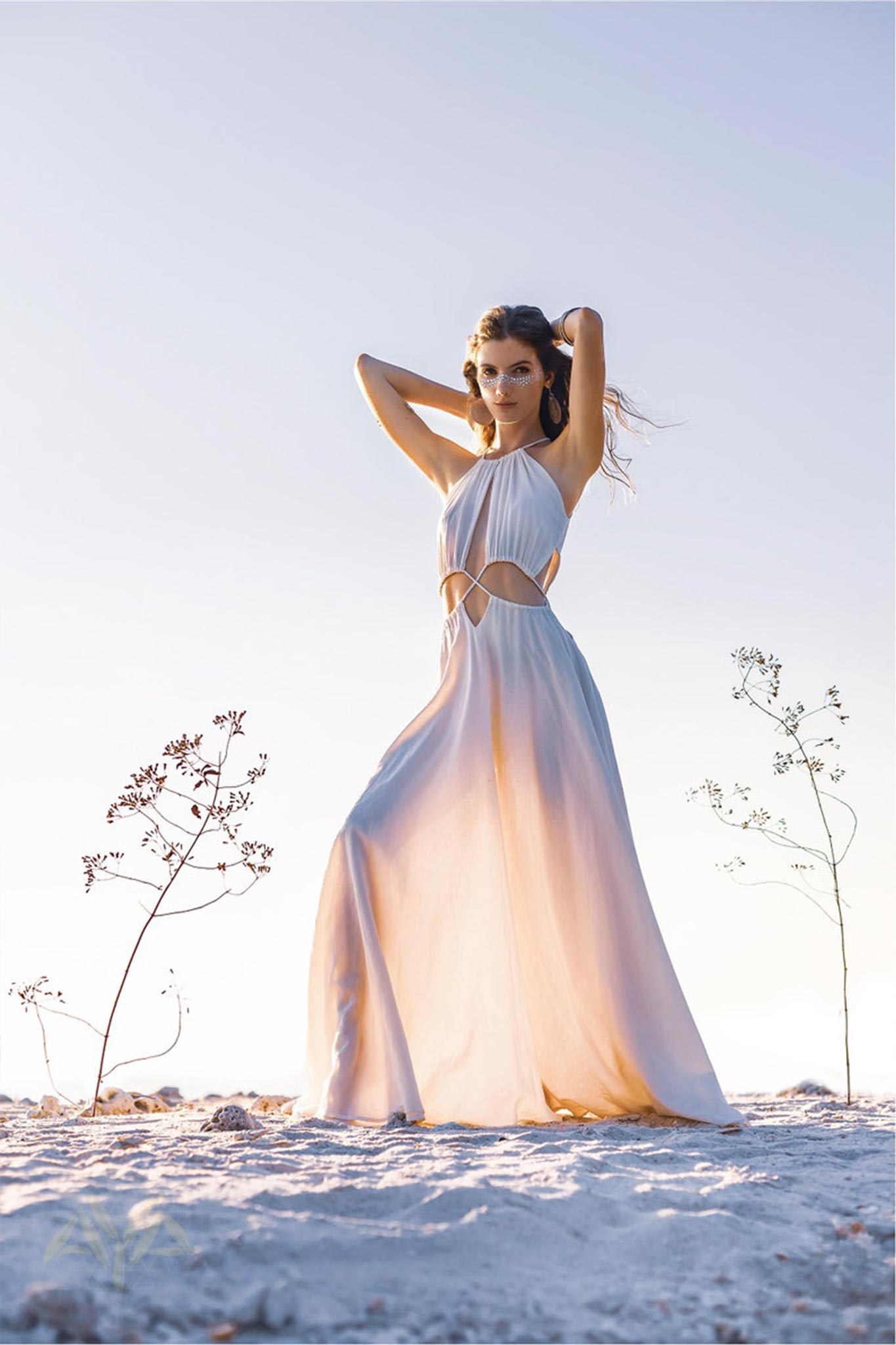 A woman stands outdoors in a Boho Wedding Dress for Women by AYA Sacred Wear, featuring open back and sides with cut-out details, crafted from organic botanically dyed fabric. Her long hair flows as she stands on a sandy surface with sparse vegetation under a clear blue sky.