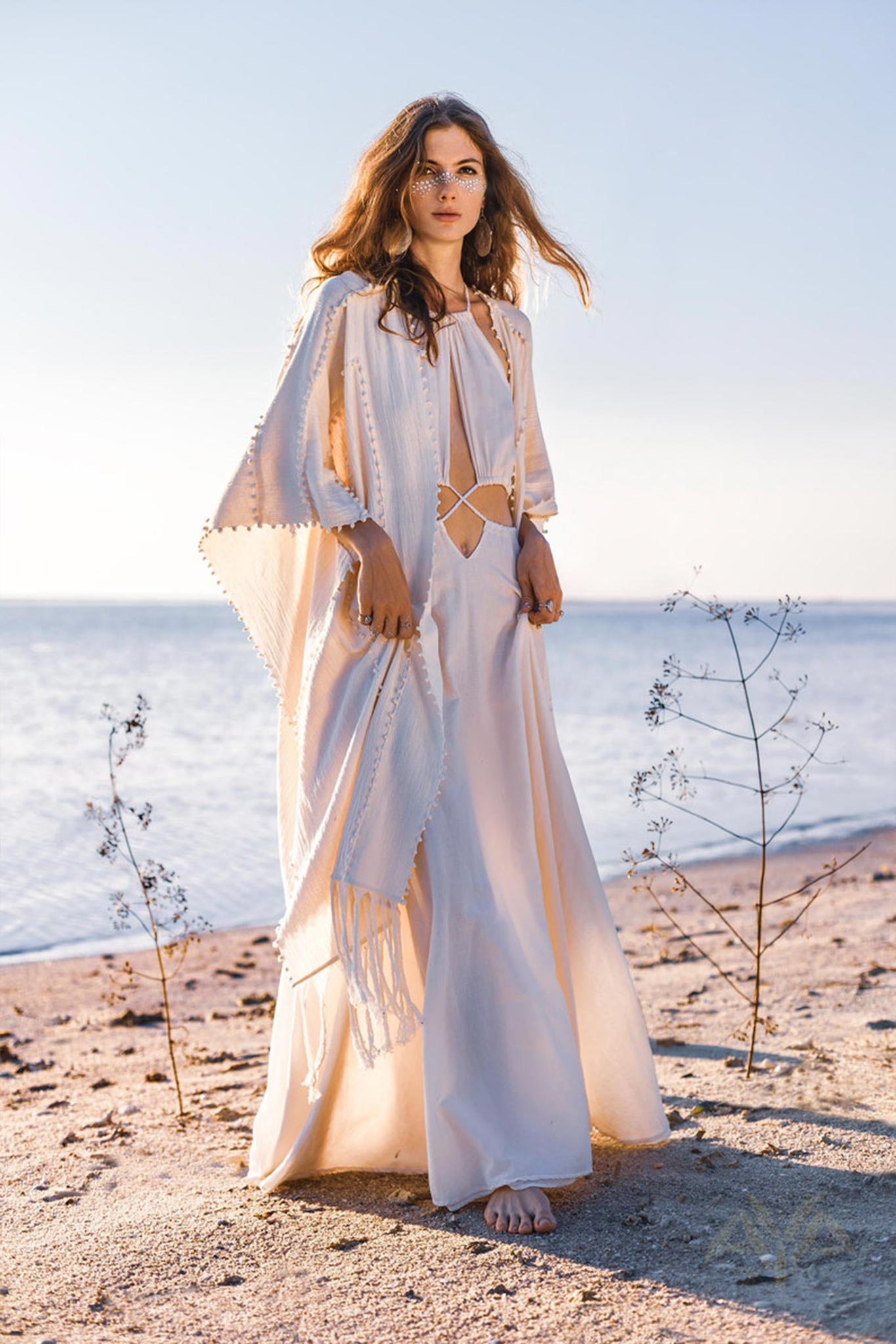 A woman stands on a beach in an AYA Sacred Wear Boho Wedding Dress for Women, featuring an open back and sides crafted from organic botanically dyed fabric. Sparse plants surround her as the ocean remains calm in the background under a clear sky, with her long hair flowing naturally.