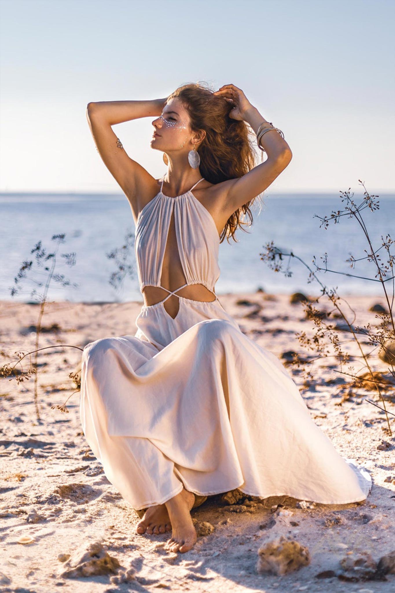 A woman wearing the AYA Sacred Wear Boho Wedding Dress with open back sides and belly design poses gracefully on a sandy beach by calm ocean waves. With her hands in her hair, she gazes upward, while wispy plants in the foreground frame her against a clear sky, enhancing the organic beauty of her attire.