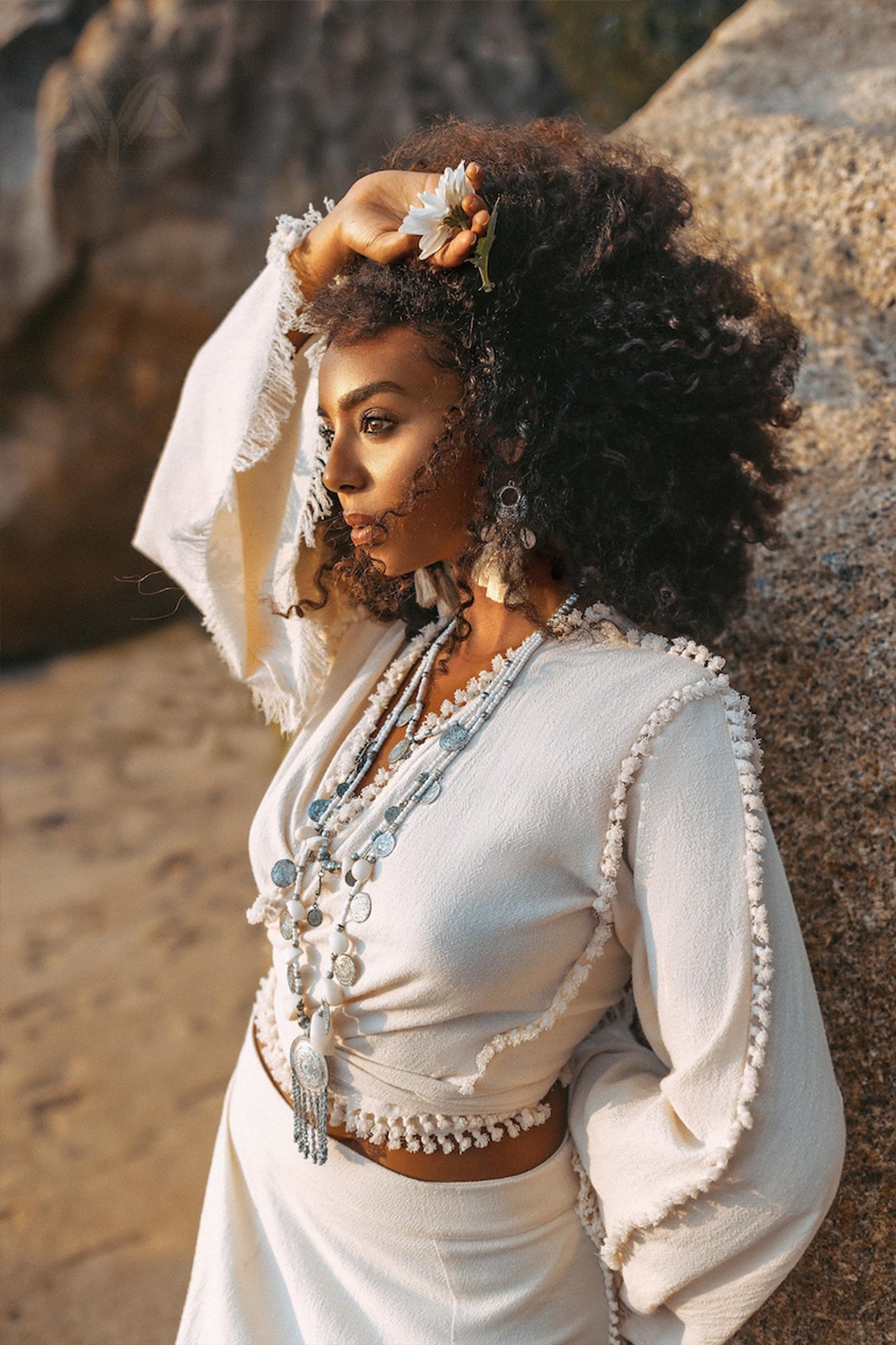 Wearing the Boho Wrap Top from AYA Sacred Wear, which features an organic cotton fabric and wide sleeves, a woman poses against a rocky backdrop with a flower nestled in her curly hair. Adorned with layered necklaces, she gazes into the distance amidst a tranquil beach setting.