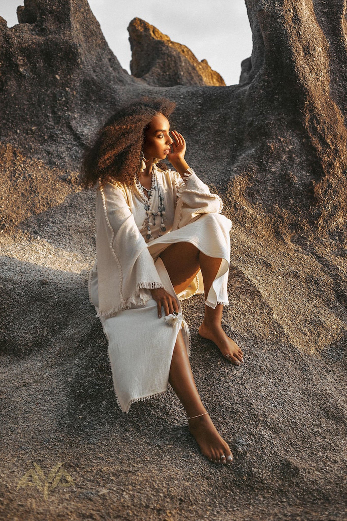 A woman in a Boho Wrap Top by AYA Sacred Wear, featuring organic wide sleeves, sits barefoot on rocky terrain, surrounded by large stone formations. Her curly hair gently blows in the wind as she gazes thoughtfully into the distance. The scene is serene under soft lighting.