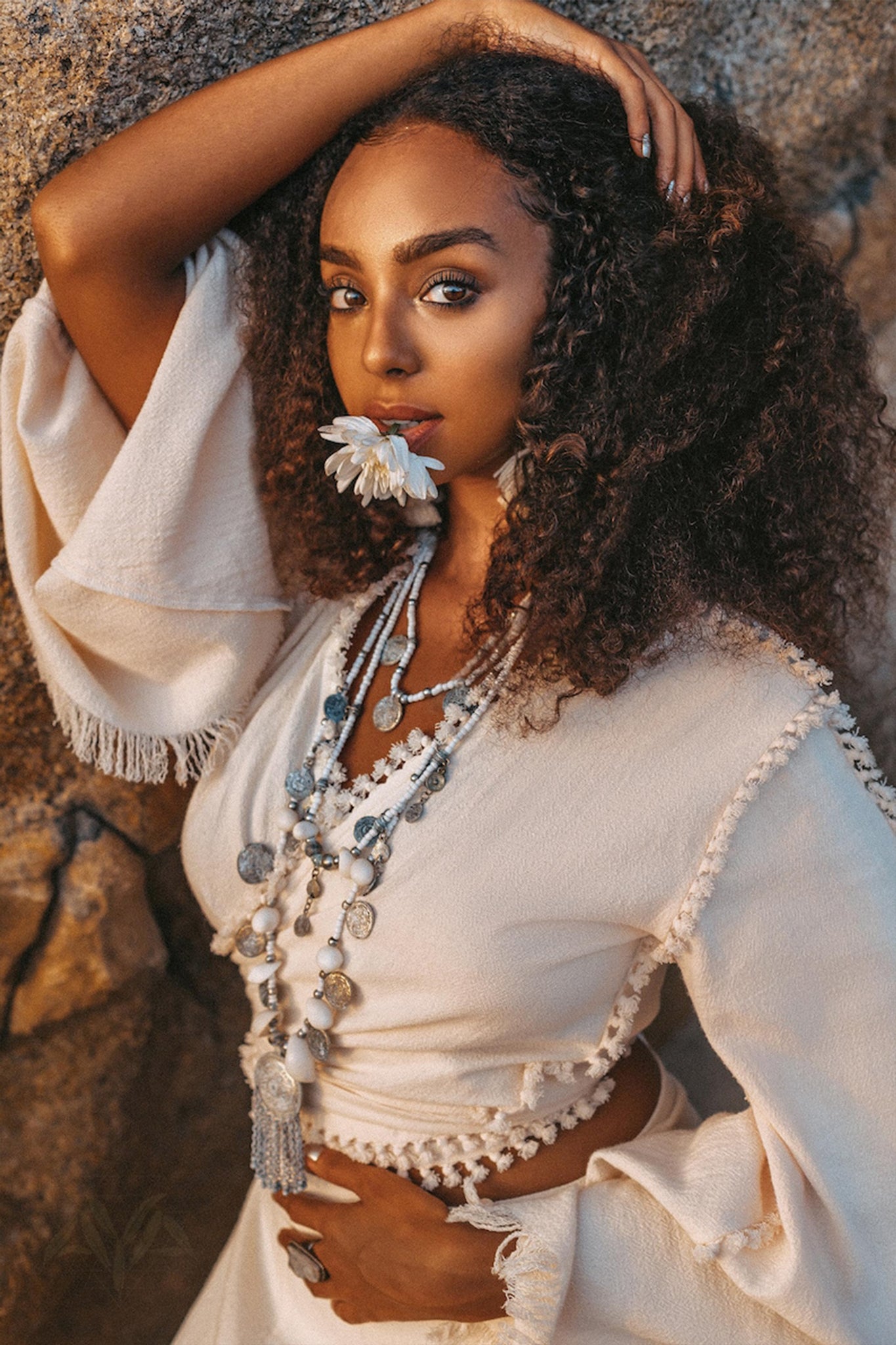 A woman with curly hair strikes a pose against a rock, dressed in the AYA Sacred Wear Boho Wrap Top • Organic Wide Sleeve Blouse • Bridal Goddess Top. She accessorizes with layered necklaces while holding a white flower to her lips and gazing at the camera, creating a serene and natural ambiance that perfectly complements her organic cotton outfit.