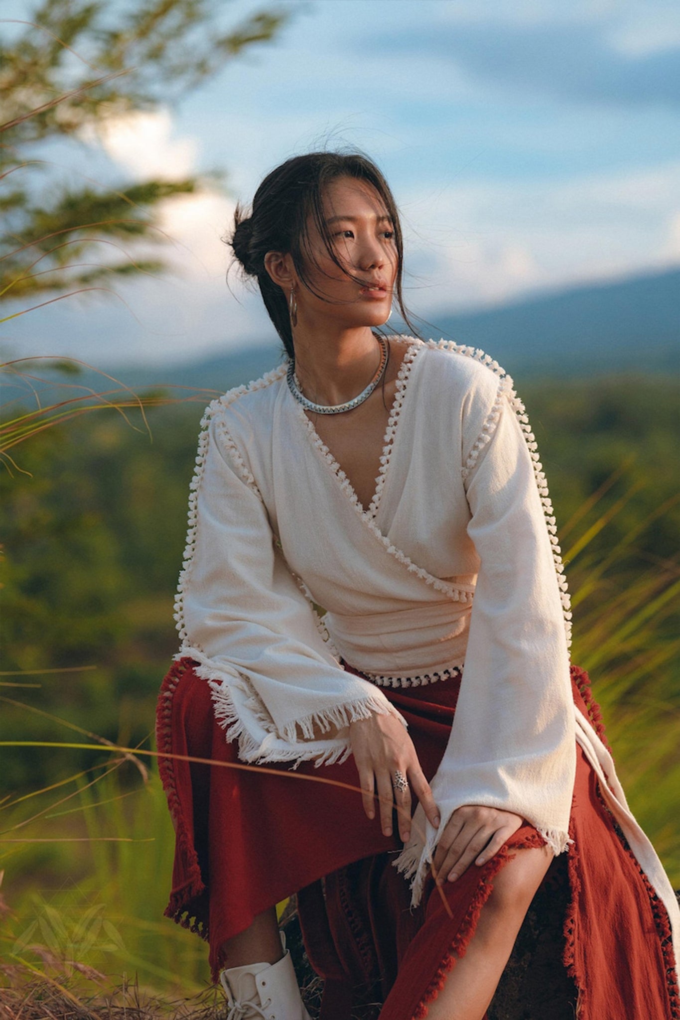 A person with long dark hair sits outdoors, dressed in the AYA Sacred Wear Boho Wrap Top adorned with ornamental trim, paired with rust-colored pants. They gaze to the side, set against a backdrop of lush green foliage and a distant mountain under a partly cloudy sky.