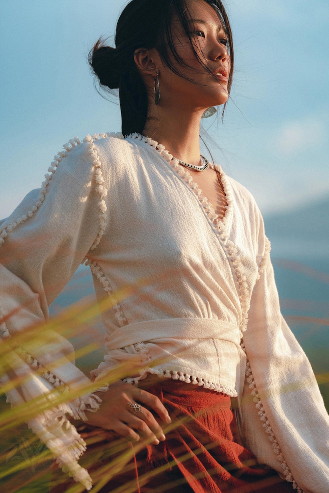 A woman stands in a sunlit field, wearing the Boho Wrap Top—an Organic Wide Sleeve Blouse by AYA Sacred Wear—and a rust-colored skirt. Her hair is styled in a bun, and she is adorned with delicate jewelry. Tall grasses frame the foreground against a clear blue sky.