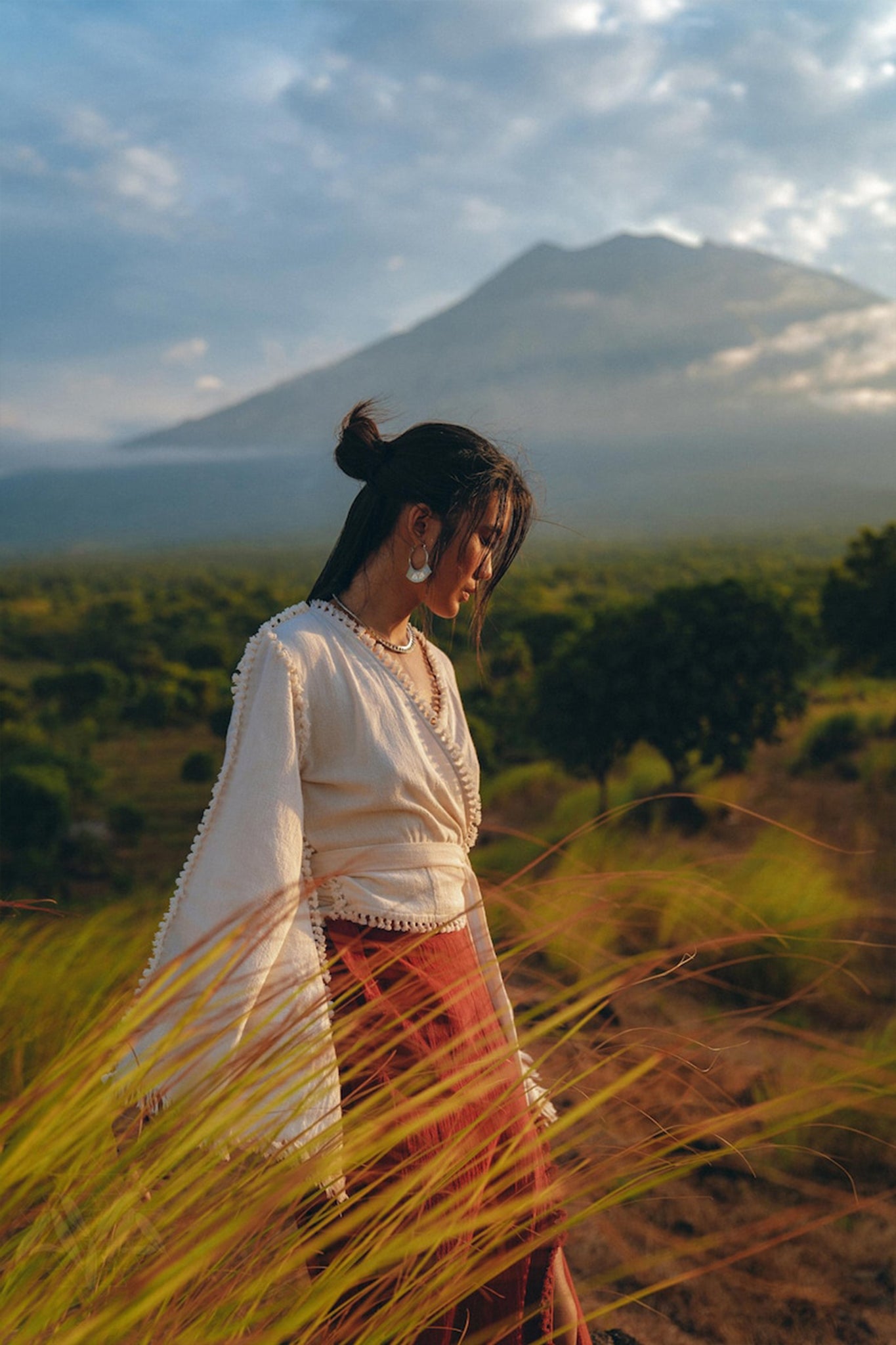 A woman stands in a field of tall grasses, dressed in the AYA Sacred Wear Boho Wrap Top paired with a red skirt. She appears contemplative, her head slightly bowed. In the background, a misty mountain rises under a blue, cloud-dappled sky.