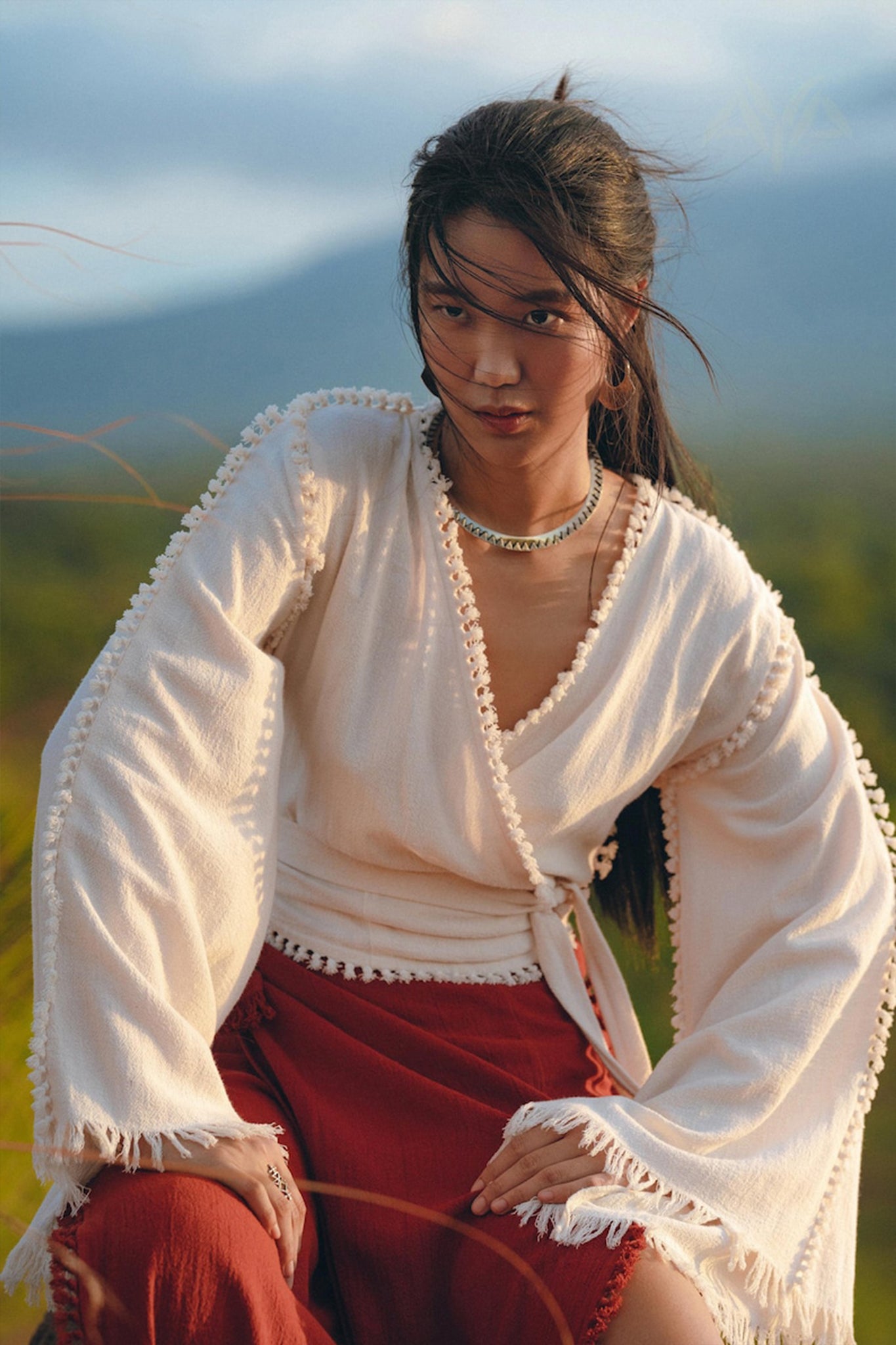 A person with long hair, dressed in AYA Sacred Wear's Boho Wrap Top, stands in a natural setting with blurred greenery and mountains in the background. The organic wide sleeve blouse exudes effortless charm as the person gazes intensely at the camera, paired beautifully with red pants.