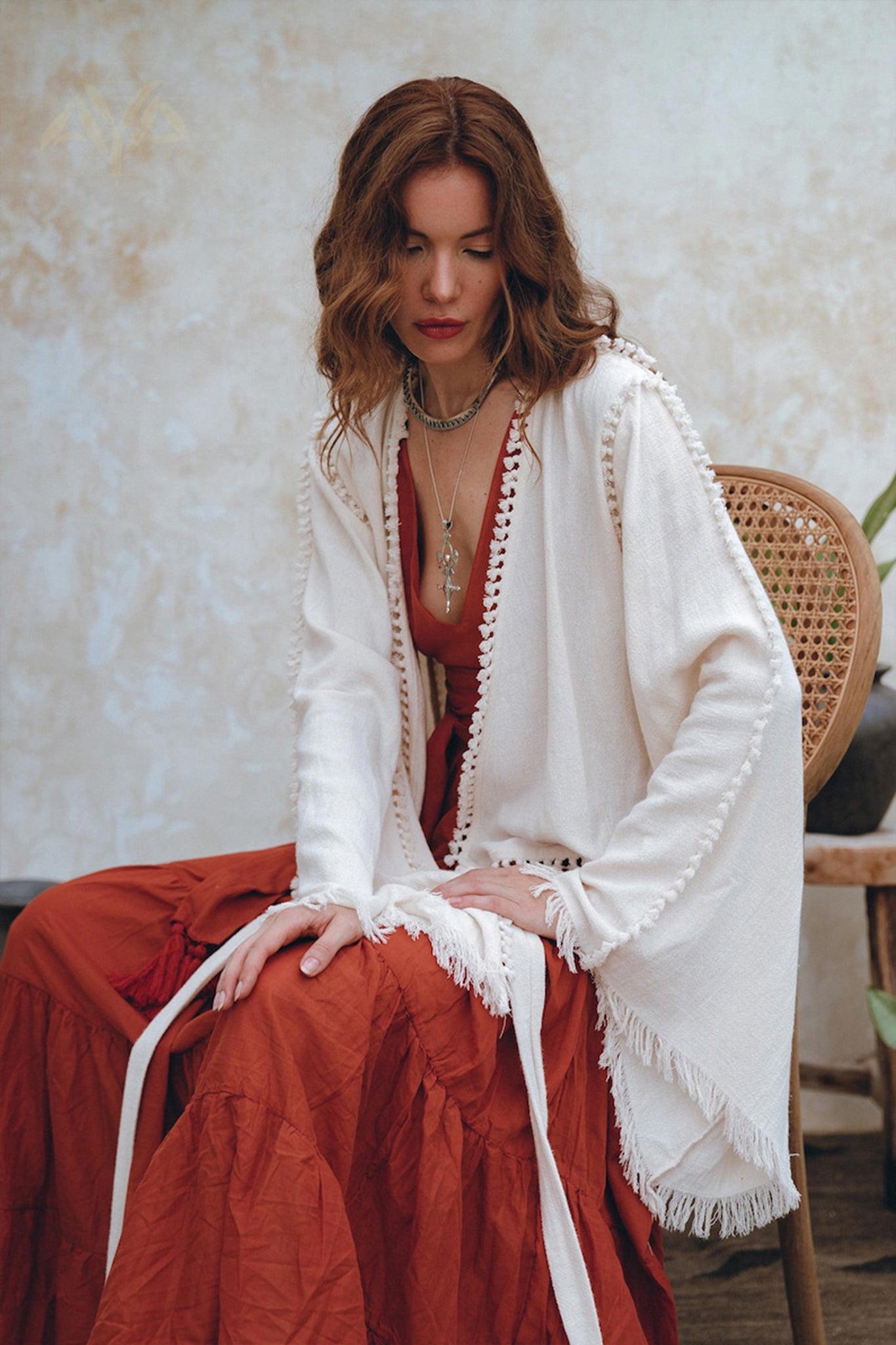 A woman with wavy hair sits on a chair in a long, burnt orange dress, complemented by a textured white cardigan elegantly crafted from organic cotton. She looks down, her look completed by layered necklaces that enhance her boho style against neutral-toned walls. She's wearing the Boho Wrap Top by AYA Sacred Wear.