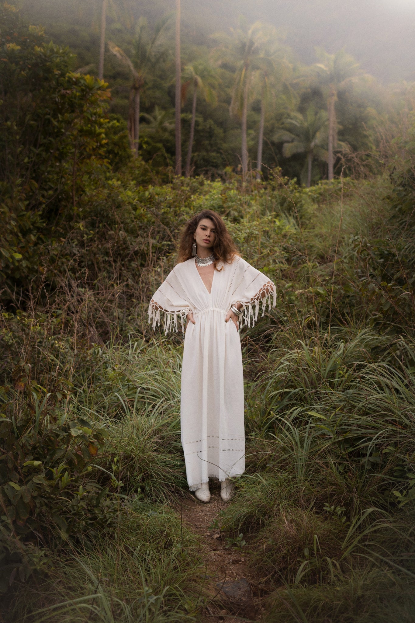 A woman in an Off-White Boho Dress by AYA Sacred Wear stands on a narrow forest path. Her long brown hair and pearl necklace complement the misty, tropical backdrop of lush greenery and tall palm trees, accentuating the elegance of her traditional handmade needlework attire.