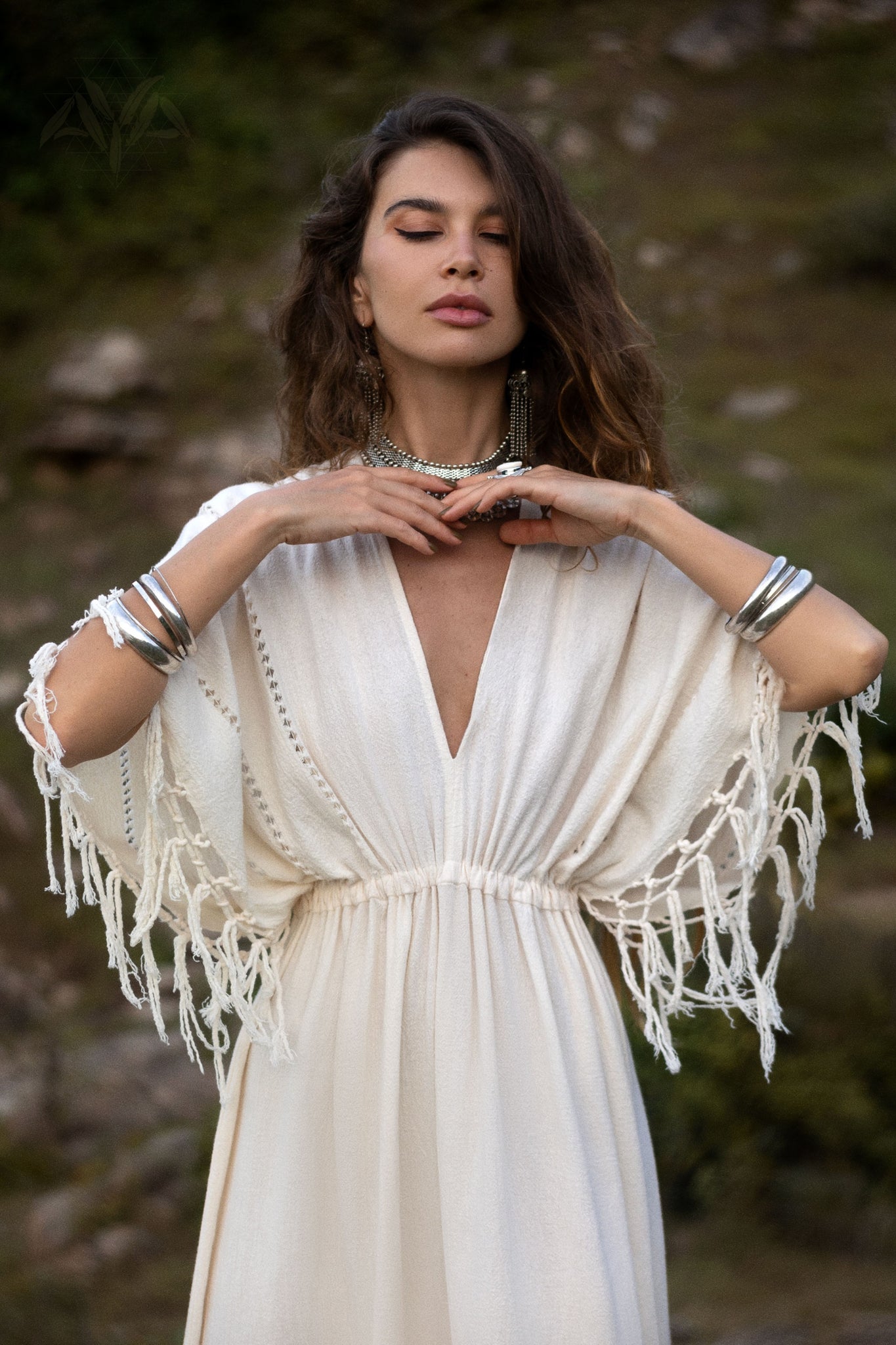 A brown-haired woman, eyes closed, stands outside in an off-white AYA Sacred Wear Boho Goddess Dress with fringed sleeves. She wears accessories like a necklace, earrings, and silver bracelets against a blurred natural backdrop enhancing the traditional handmade needlework of her attire.