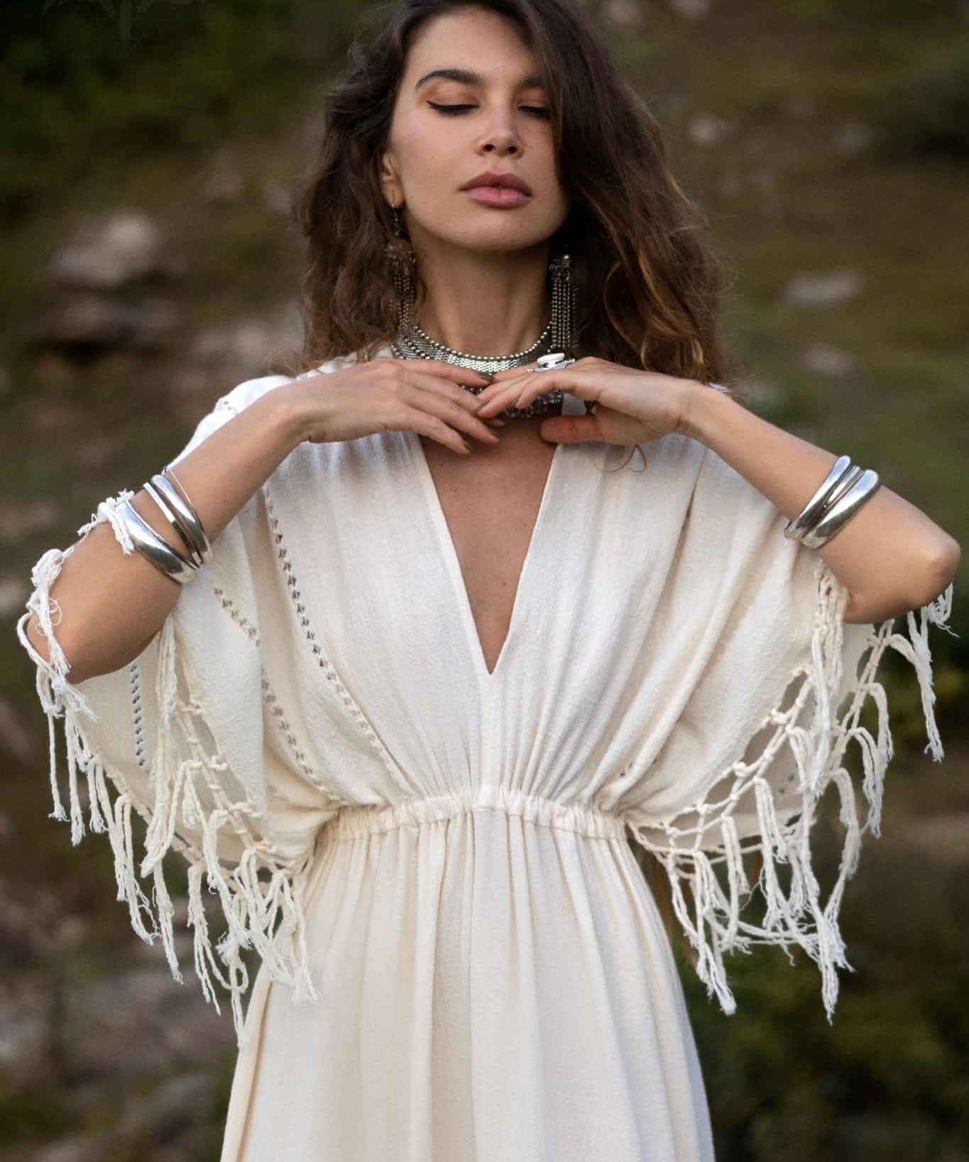 A woman with long, wavy hair stands with closed eyes against a natural backdrop, wearing the Off-White Boho Goddess Dress from AYA Sacred Wear, featuring fringe details and handmade needlework. She pairs it perfectly with silver bracelets, a statement necklace, and earrings.