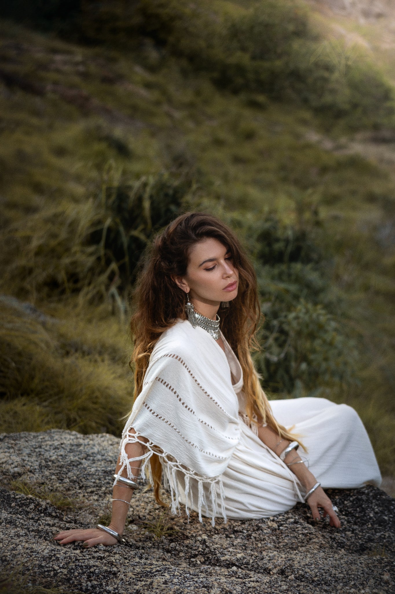 A woman with long hair sits on rocks in a grassy area, gracefully adorned in the AYA Sacred Wear Off-White Boho Dress, featuring traditional handmade needlework. She wears silver earrings and bracelets, eyes closed, radiating serenity against the blurred backdrop.