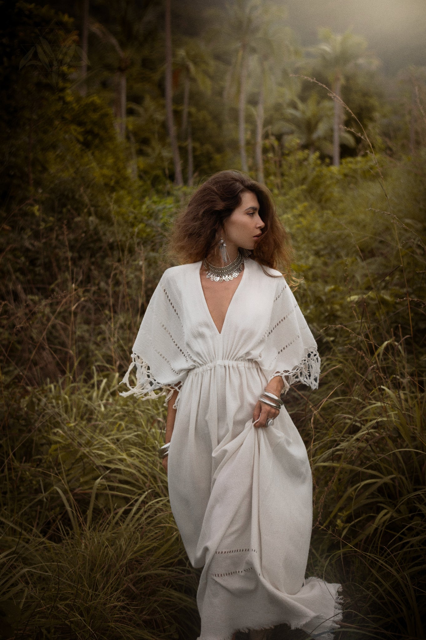 In a dreamy setting of lush greenery with tall grass and palm trees, a woman stands in an Off-White Boho Dress by AYA Sacred Wear. Her long, wavy hair complements her handmade statement jewelry against the misty backdrop.