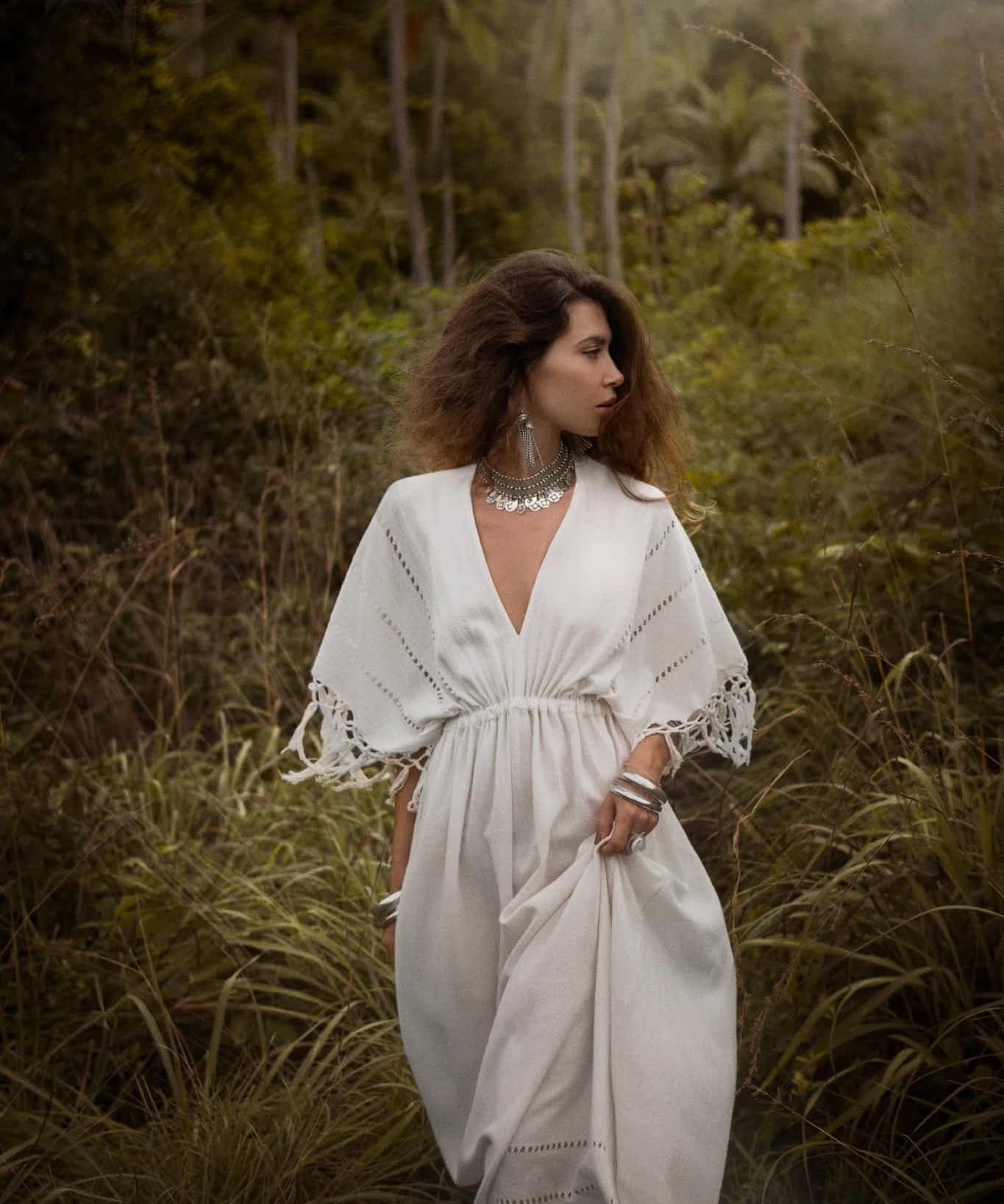 In a lush green forest, a woman stands wearing an AYA Sacred Wear Off-White Boho Dress adorned with traditional handmade needlework. Her long brown hair and silver jewelry accentuate the misty, serene atmosphere of tall trees surrounding her.